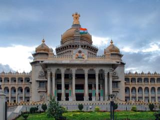 Facade of the Vidhana Soudha building in Bengaluru, with an Indian flag on its central dome under a cloudy sky