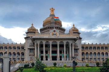 Facade of the Vidhana Soudha building in Bengaluru, with an Indian flag on its central dome under a cloudy sky