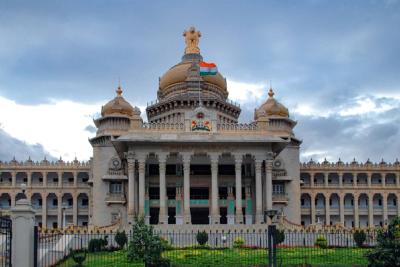 Facade of the Vidhana Soudha building in Bengaluru, with an Indian flag on its central dome under a cloudy sky