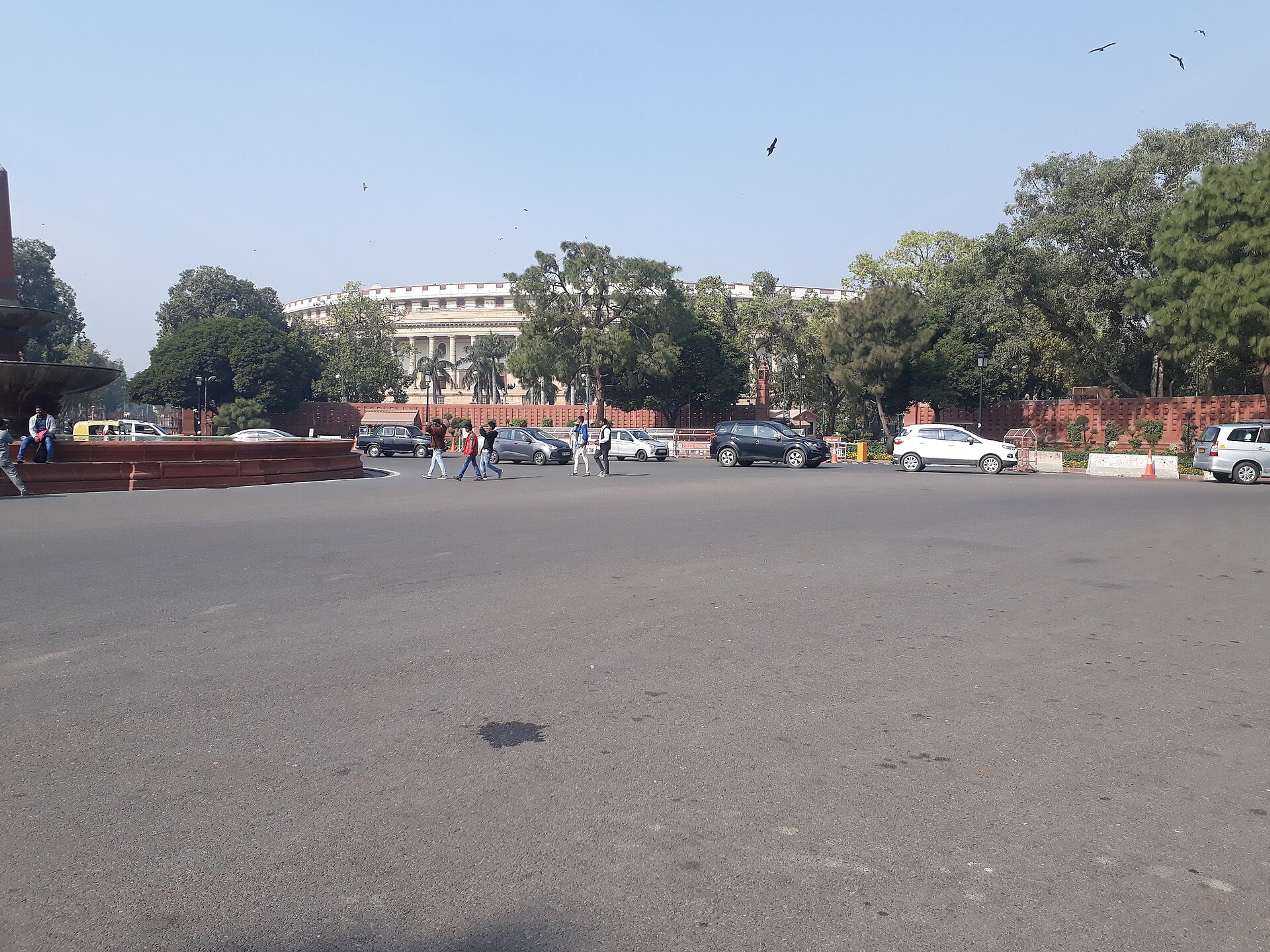 Pedestrians and parked cars on a wide road in front of the Parliament building in New Delhi, with trees and a fountain.