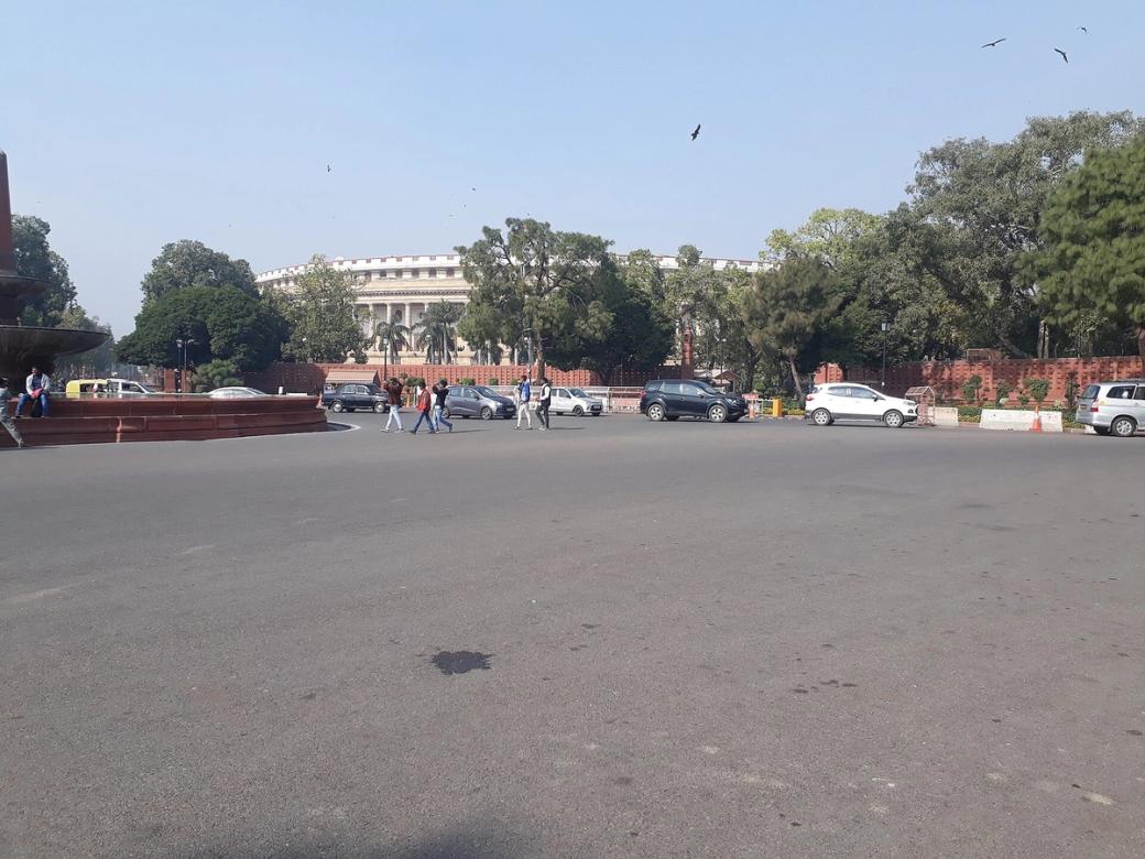 Pedestrians and parked cars on a wide road in front of the Parliament building in New Delhi, with trees and a fountain.