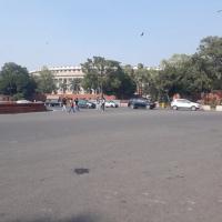 Pedestrians and parked cars on a wide road in front of the Parliament building in New Delhi, with trees and a fountain.