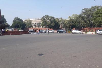 Pedestrians and parked cars on a wide road in front of the Parliament building in New Delhi, with trees and a fountain.