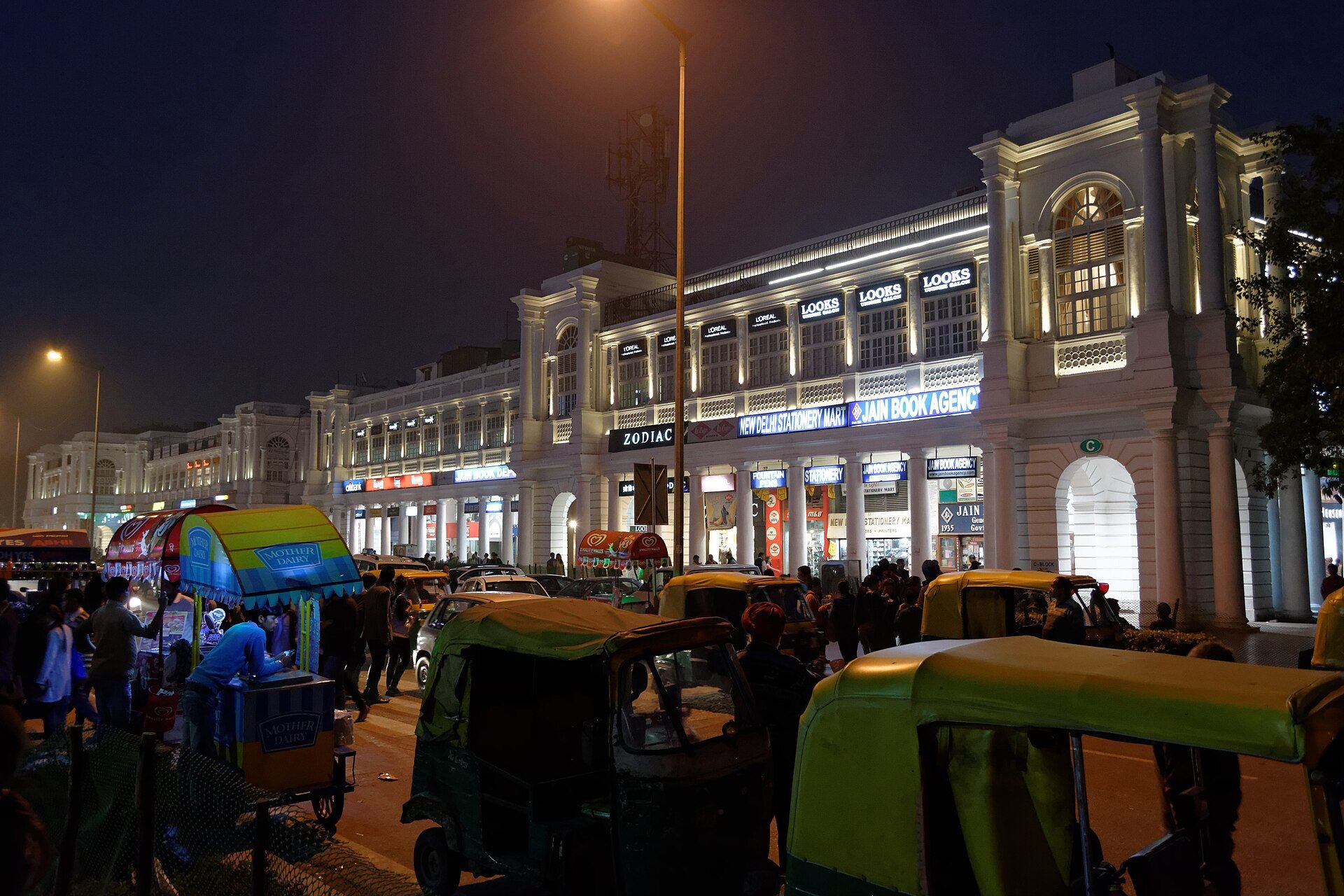Lit colonnaded buildings at Connaught Place at night, with auto-rickshaws, a street food cart and people in the foreground