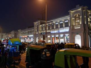 Lit colonnaded buildings at Connaught Place at night, with auto-rickshaws, a street food cart and people in the foreground