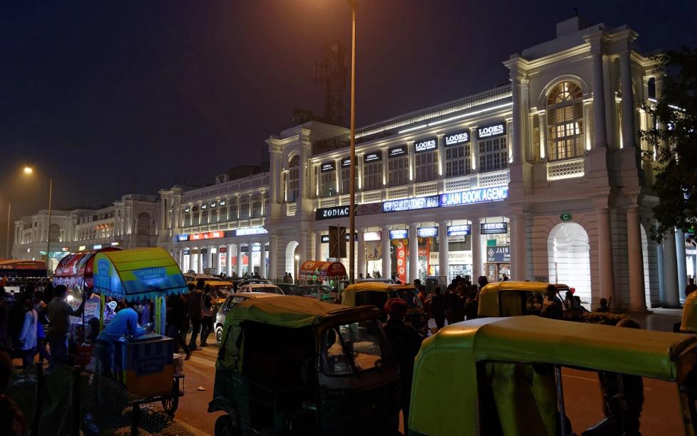 Lit colonnaded buildings at Connaught Place at night, with auto-rickshaws, a street food cart and people in the foreground