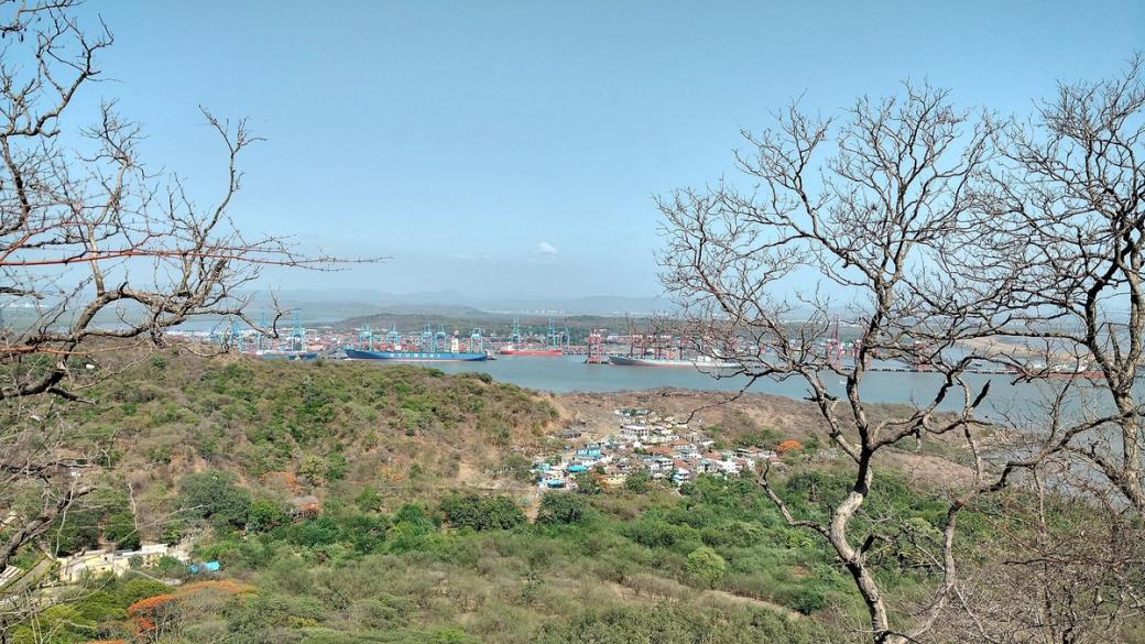 Container port with cranes and ships across water, seen from a hillside above a village of houses.