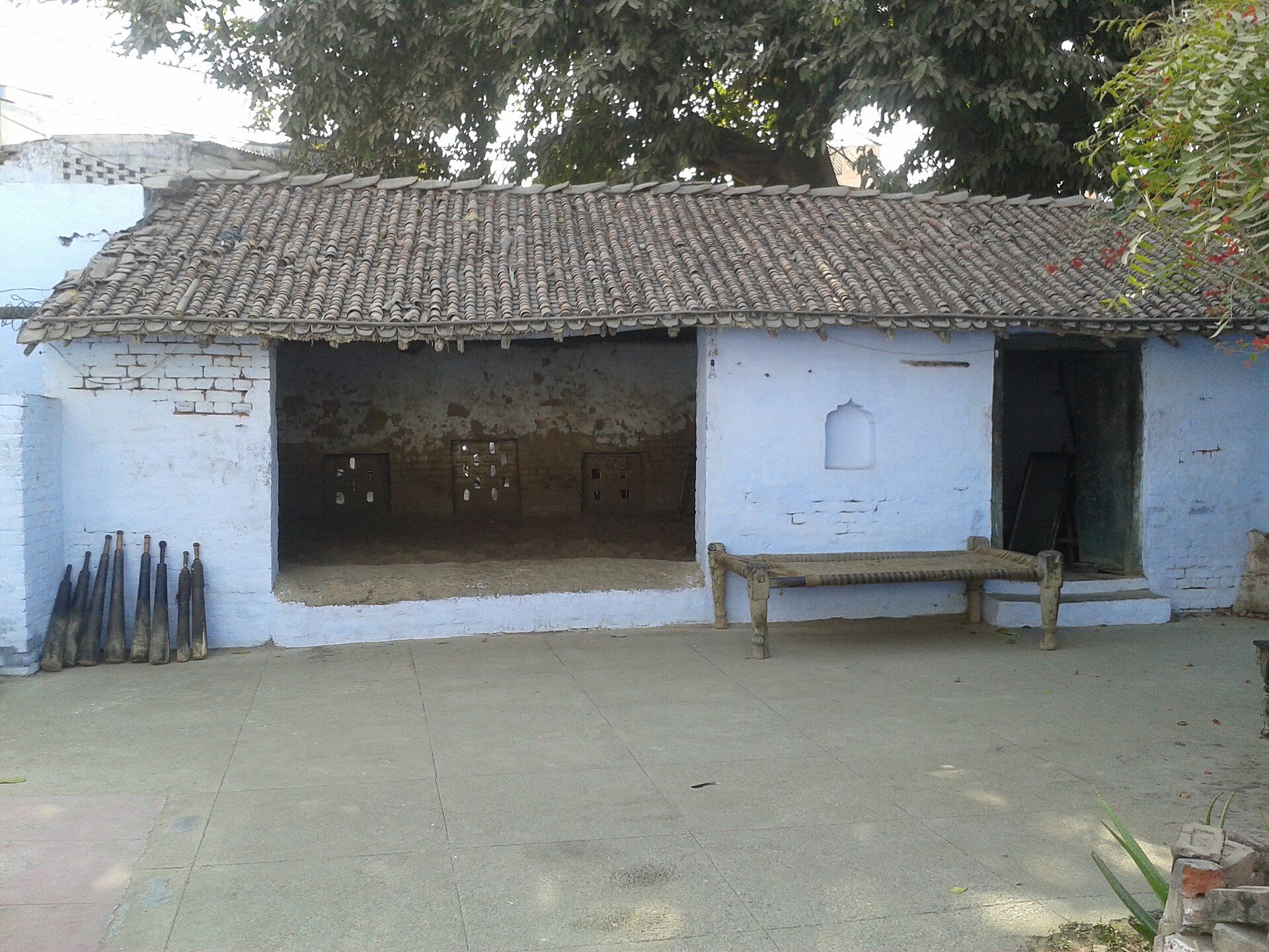 Whitewashed tiled-roof building with an open earthen-floored hall, a wooden cot and wooden clubs against the wall