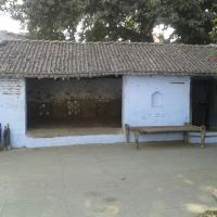 Whitewashed tiled-roof building with an open earthen-floored hall, a wooden cot and wooden clubs against the wall