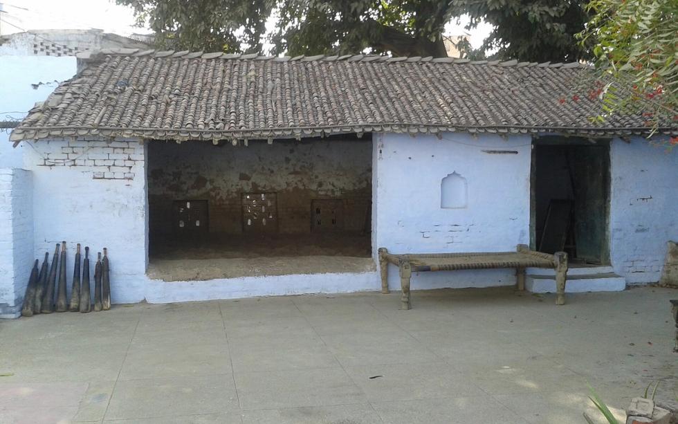 Whitewashed tiled-roof building with an open earthen-floored hall, a wooden cot and wooden clubs against the wall