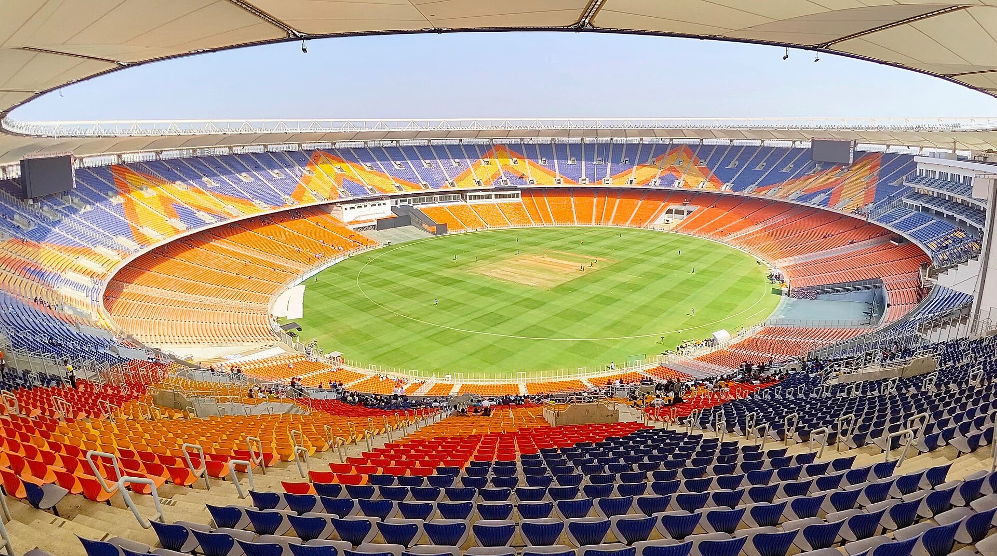Wide view of a cricket stadium with orange and blue seats around a green field under a clear sky.