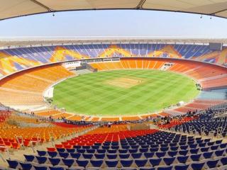 Wide view of a cricket stadium with orange and blue seats around a green field under a clear sky.