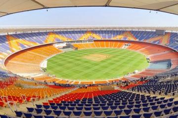 Wide view of a cricket stadium with orange and blue seats around a green field under a clear sky.