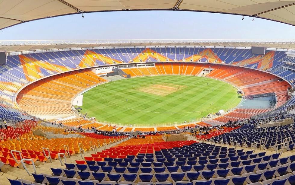 Wide view of a cricket stadium with orange and blue seats around a green field under a clear sky.