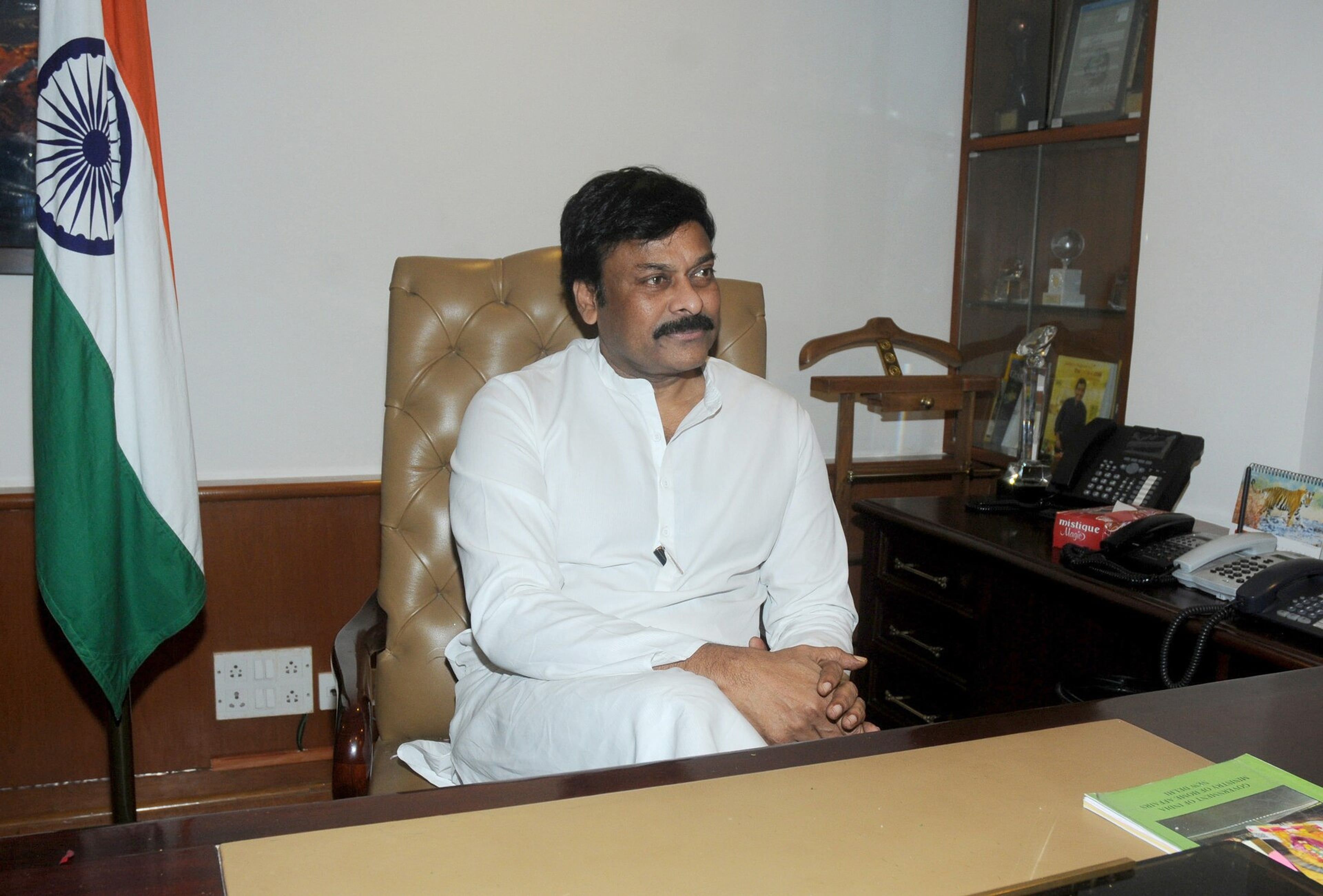 Man in a white kurta seated in a leather chair before an Indian flag in an office, with a desk and telephones beside him.