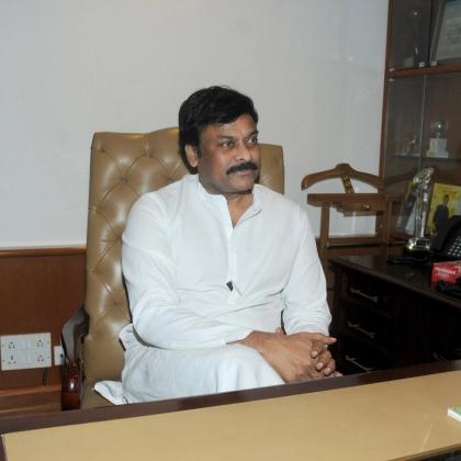 Man in a white kurta seated in a leather chair before an Indian flag in an office, with a desk and telephones beside him.