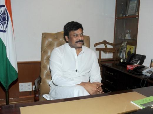 Man in a white kurta seated in a leather chair before an Indian flag in an office, with a desk and telephones beside him.