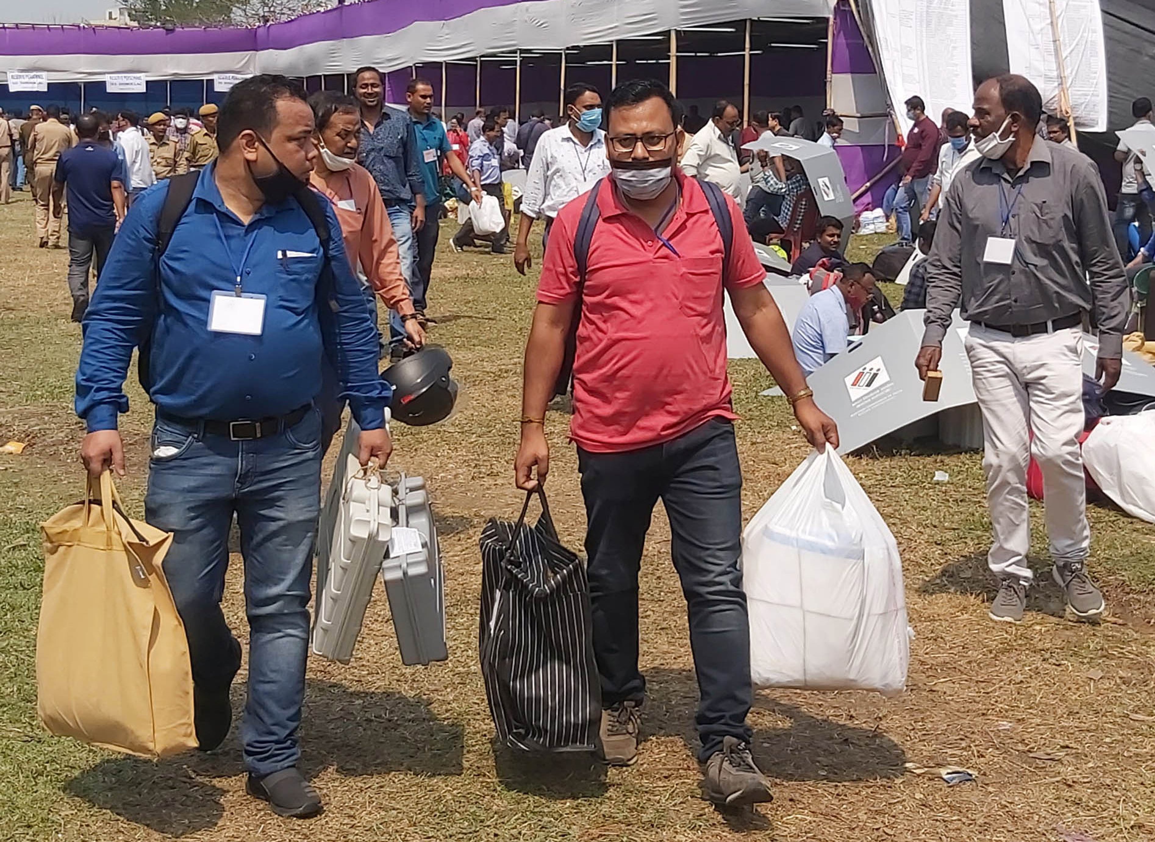 Officials in face masks carry voting machines and bags across a grassy field past a large tent.
