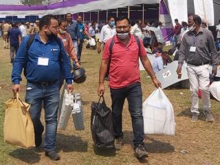 Officials in face masks carry voting machines and bags across a grassy field past a large tent.