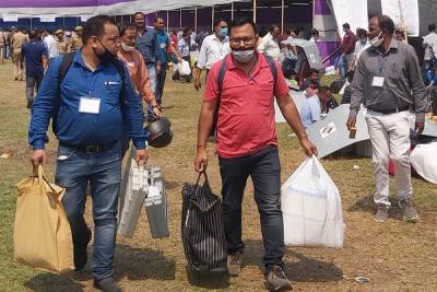 Officials in face masks carry voting machines and bags across a grassy field past a large tent.