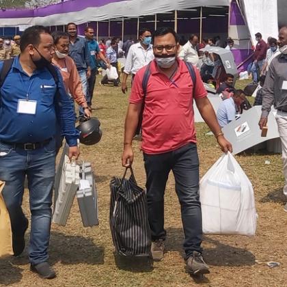 Officials in face masks carry voting machines and bags across a grassy field past a large tent.
