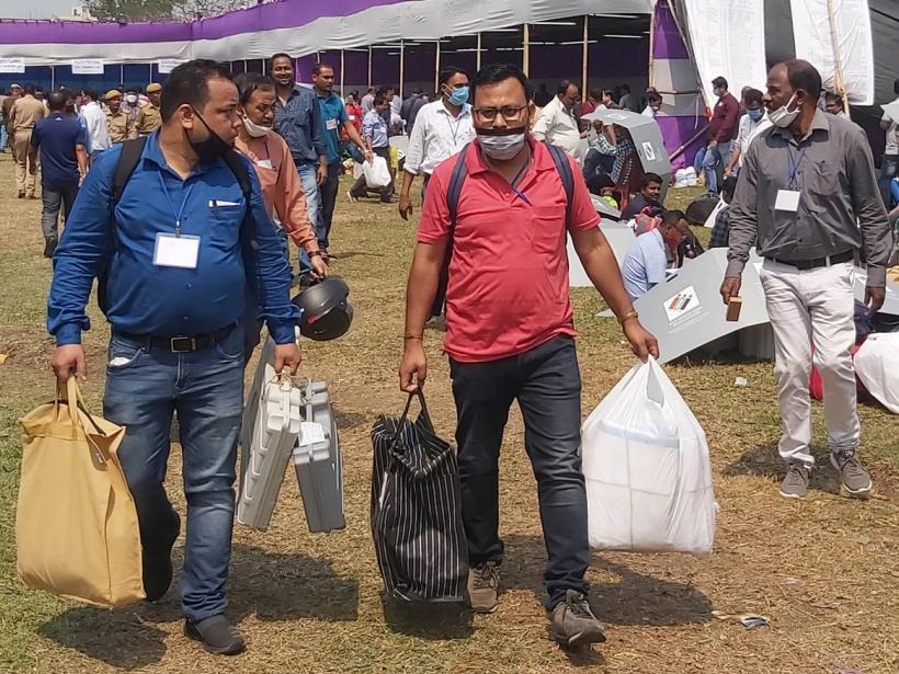 Officials in face masks carry voting machines and bags across a grassy field past a large tent.