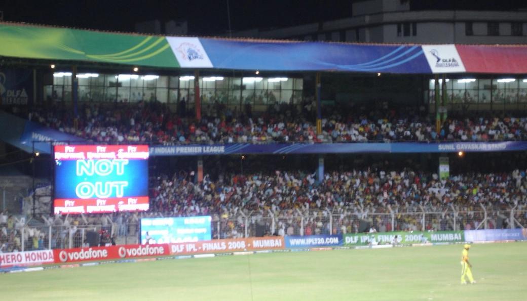 Floodlit cricket stadium at night with a NOT OUT scoreboard, a crowd in the stands, and a fielder on the grass