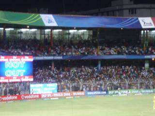 Floodlit cricket stadium at night with a NOT OUT scoreboard, a crowd in the stands, and a fielder on the grass