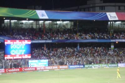 Floodlit cricket stadium at night with a NOT OUT scoreboard, a crowd in the stands, and a fielder on the grass