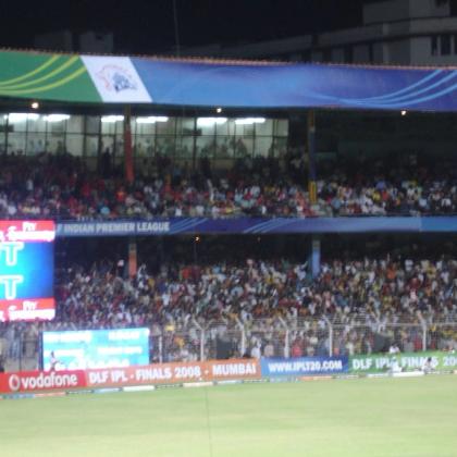 Floodlit cricket stadium at night with a NOT OUT scoreboard, a crowd in the stands, and a fielder on the grass