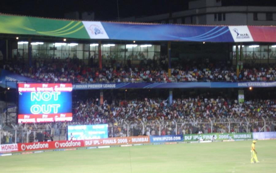 Floodlit cricket stadium at night with a NOT OUT scoreboard, a crowd in the stands, and a fielder on the grass