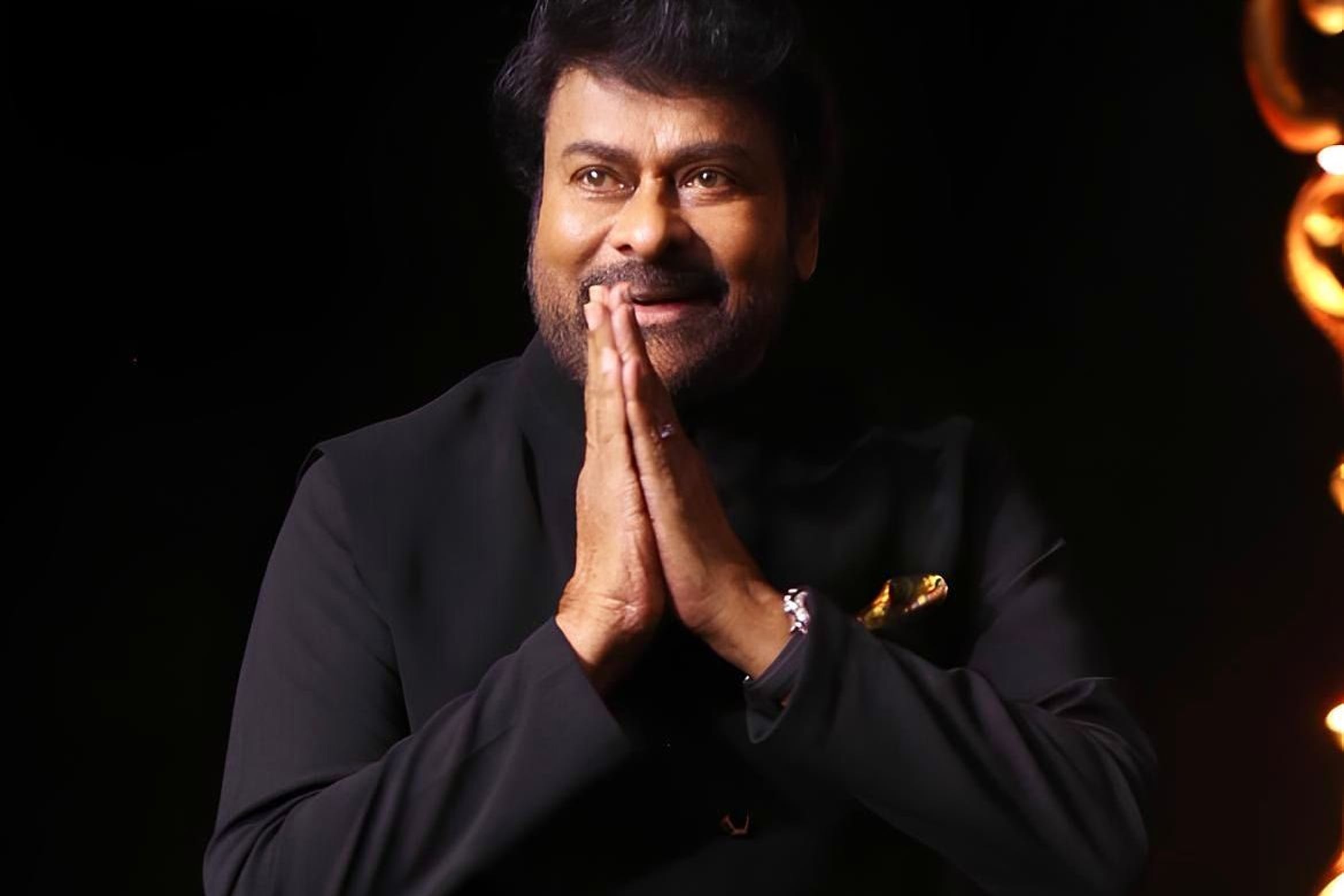 Chiranjeevi in a black bandhgala jacket, hands joined in a namaste greeting, against a dark stage background.