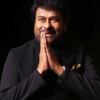 Chiranjeevi in a black bandhgala jacket, hands joined in a namaste greeting, against a dark stage background.