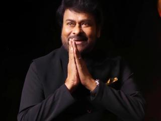 Chiranjeevi in a black bandhgala jacket, hands joined in a namaste greeting, against a dark stage background.