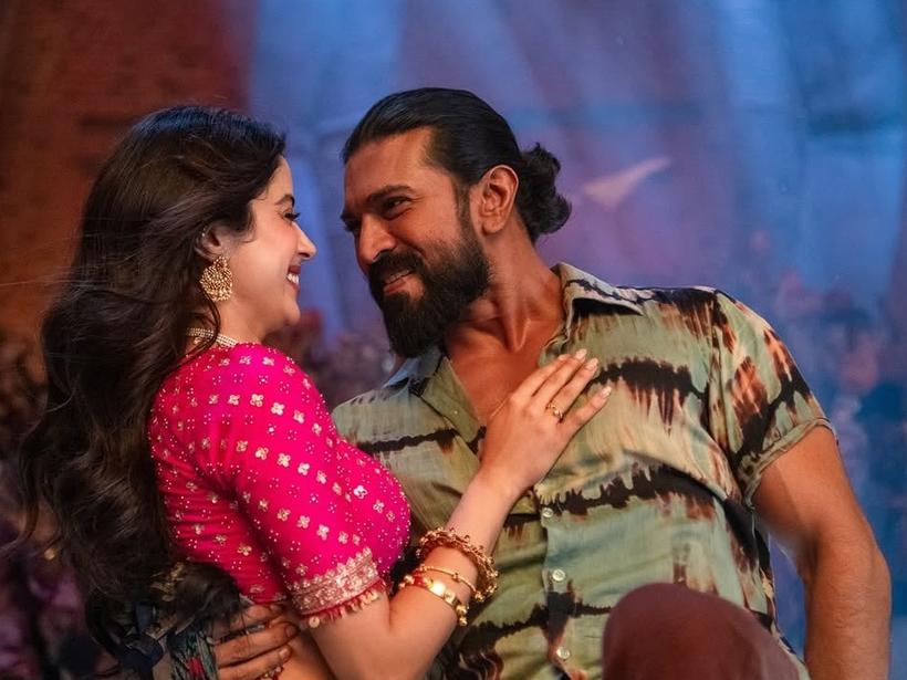 Janhvi Kapoor, in a pink blouse and gold jewellery, and Ram Charan, in a tie-dye shirt, smile at each other in a song from Peddi
