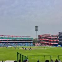 Wide view of Feroz Shah Kotla stadium with players on the field, stands with advertising banners and a floodlight tower