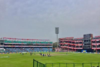 Wide view of Feroz Shah Kotla stadium with players on the field, stands with advertising banners and a floodlight tower