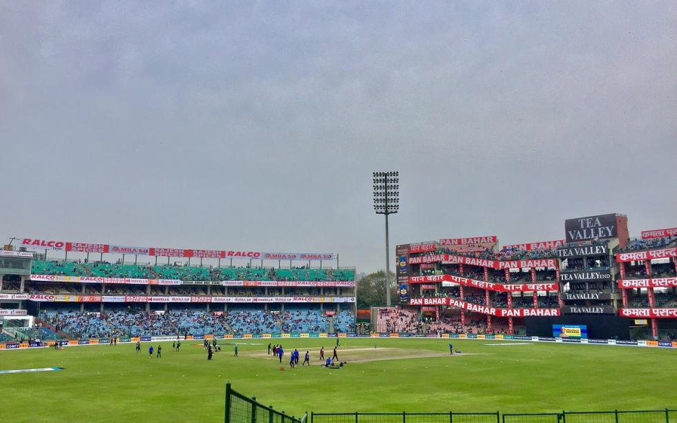 Wide view of Feroz Shah Kotla stadium with players on the field, stands with advertising banners and a floodlight tower