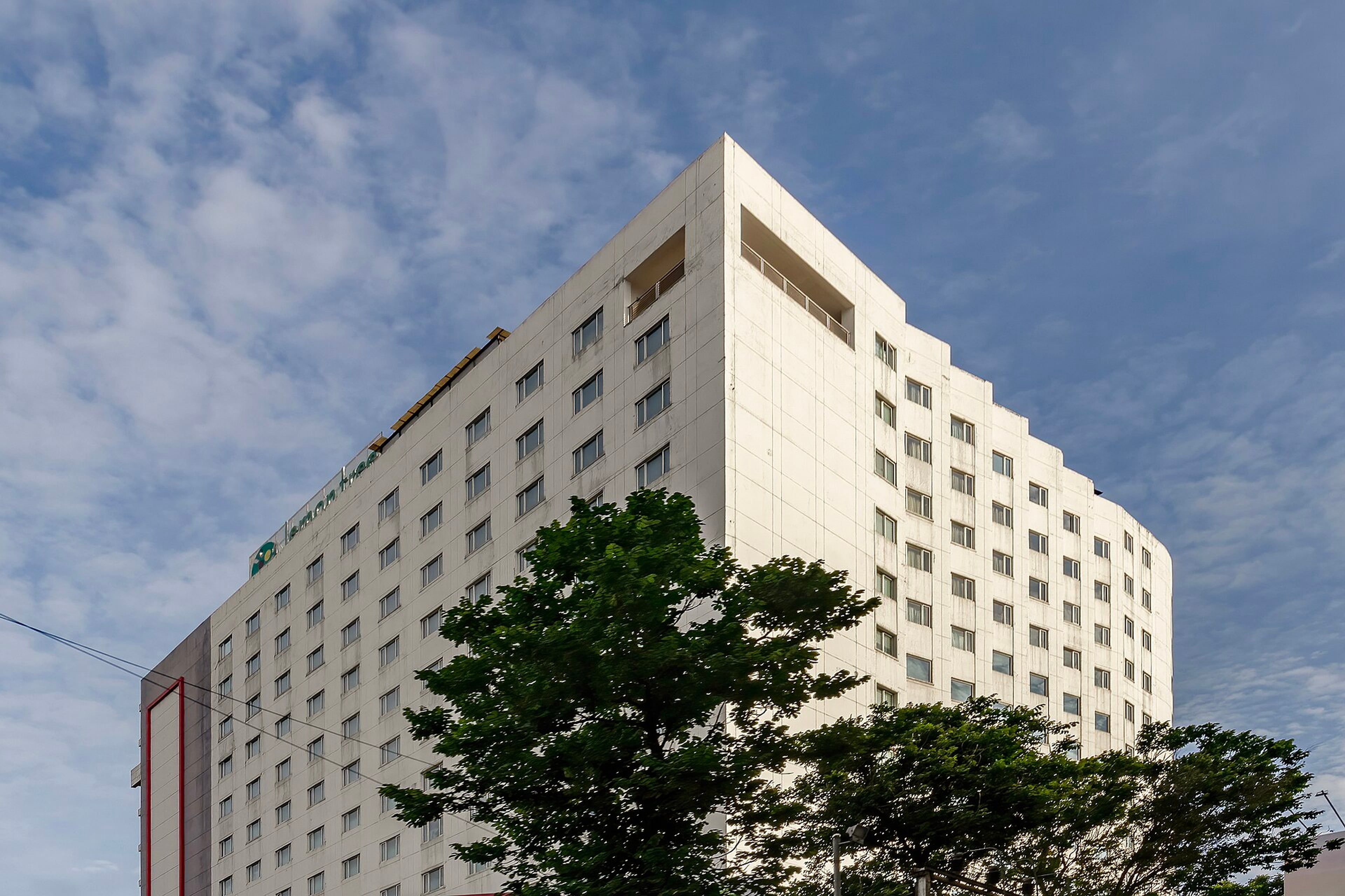Tall white hotel tower with rows of windows, trees in front and a blue sky with clouds behind it