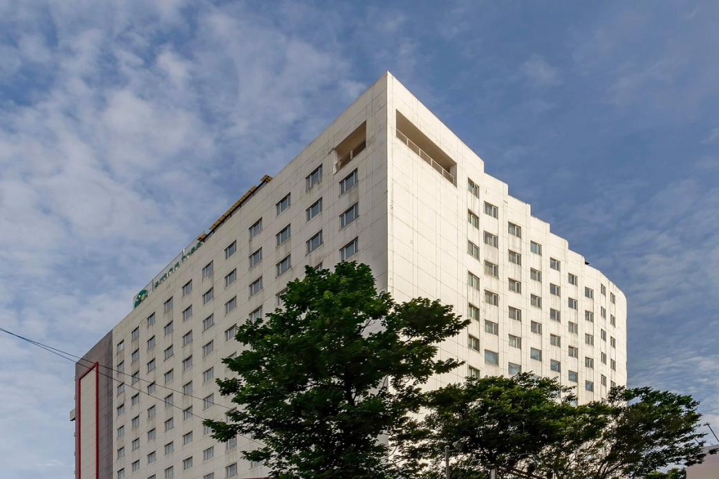 Tall white hotel tower with rows of windows, trees in front and a blue sky with clouds behind it