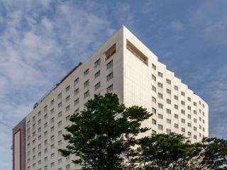 Tall white hotel tower with rows of windows, trees in front and a blue sky with clouds behind it