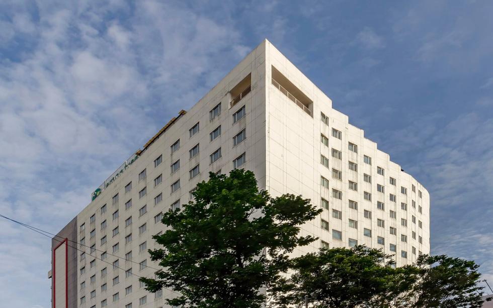 Tall white hotel tower with rows of windows, trees in front and a blue sky with clouds behind it