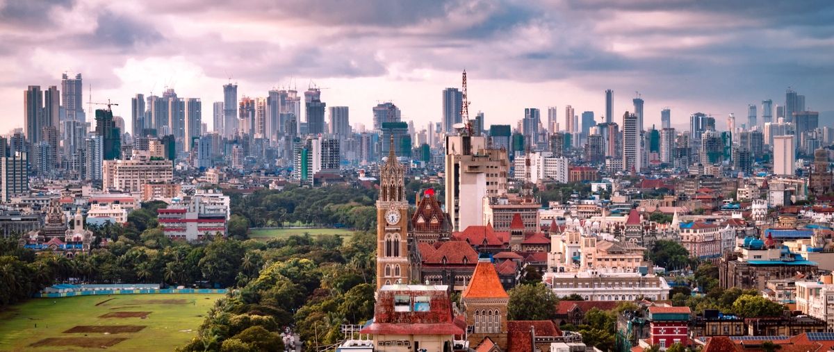 Mumbai cityscape with a clock tower, red-tiled roofs and a green field in front of distant high-rise towers