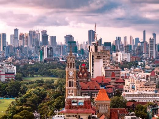 Mumbai cityscape with a clock tower, red-tiled roofs and a green field in front of distant high-rise towers