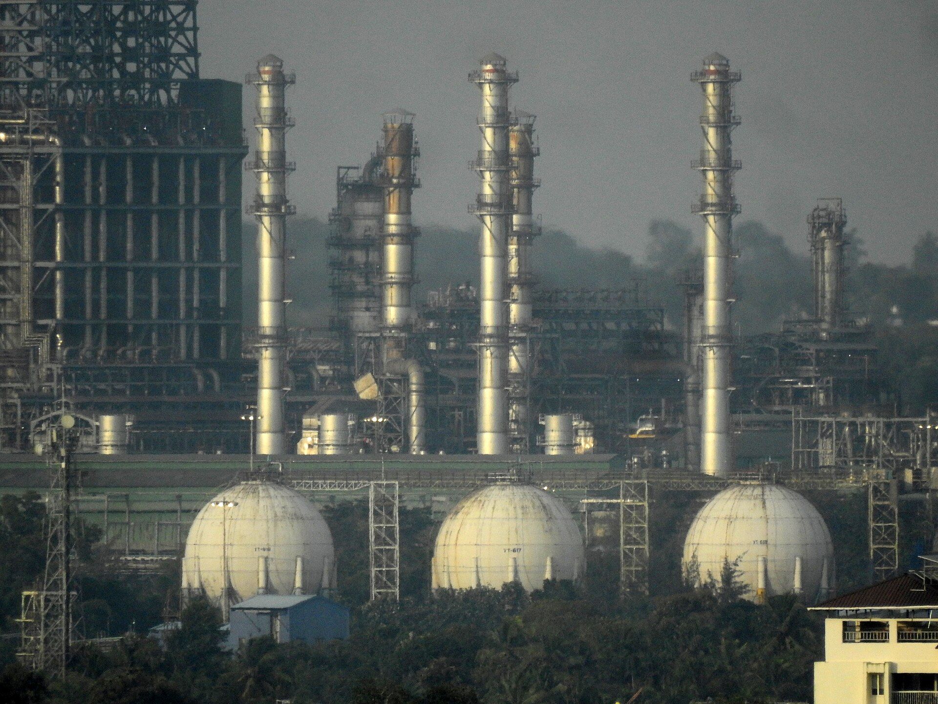 Tall refinery columns and three white spherical storage tanks among processing structures and trees