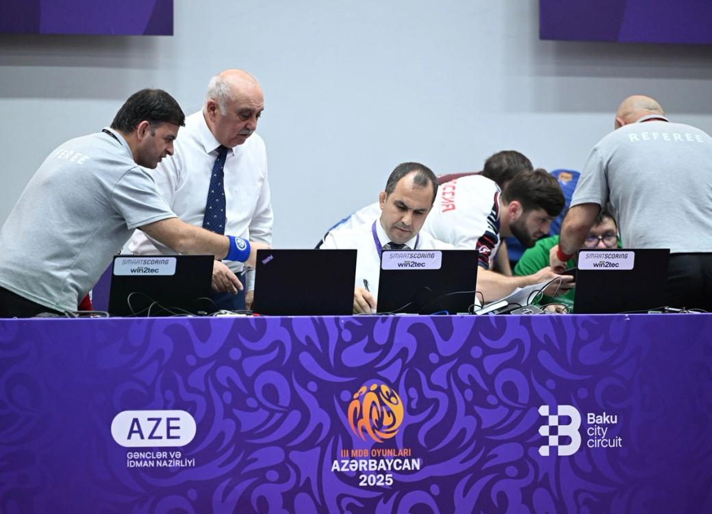 Officials and referees working at laptop scoring tables during a wrestling event, with purple event branding.