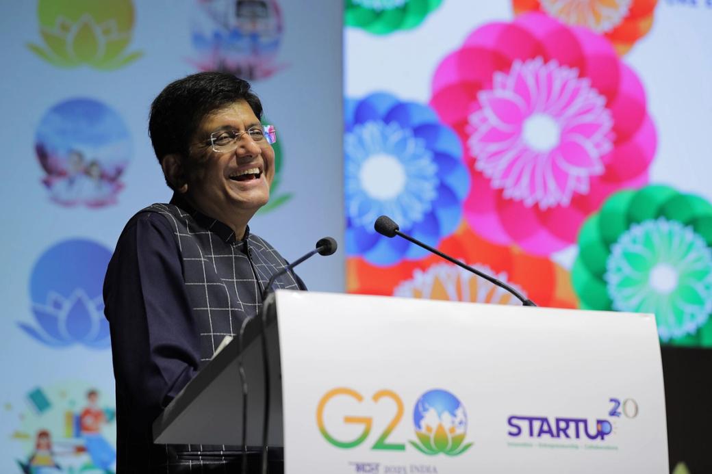 Piyush Goyal laughing as he speaks at a podium with G20 Startup20 branding, in front of colourful floral screens.