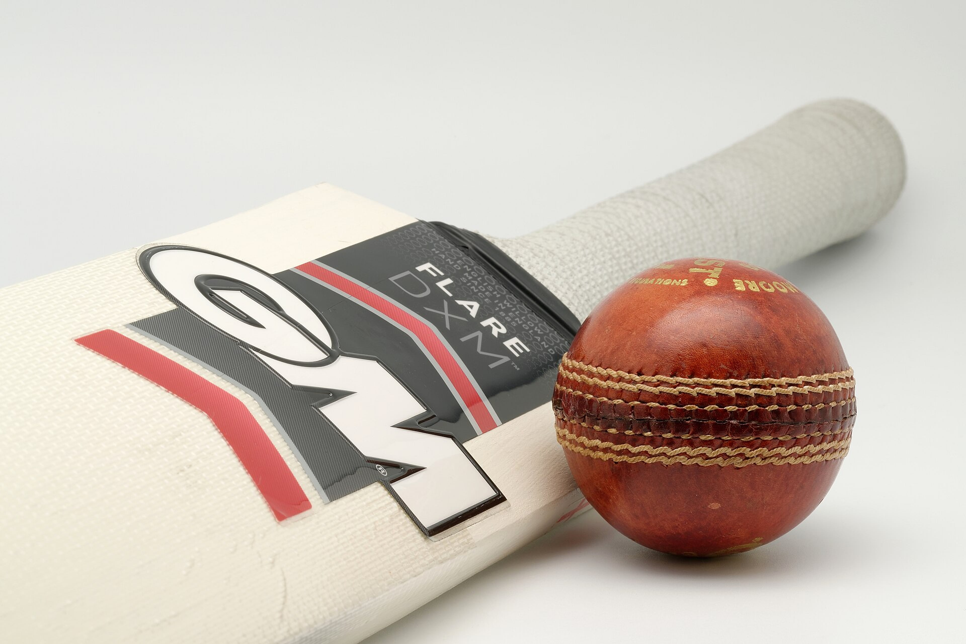 A cream cricket bat with a dark Flare DXM label and a red leather cricket ball on a plain light background.