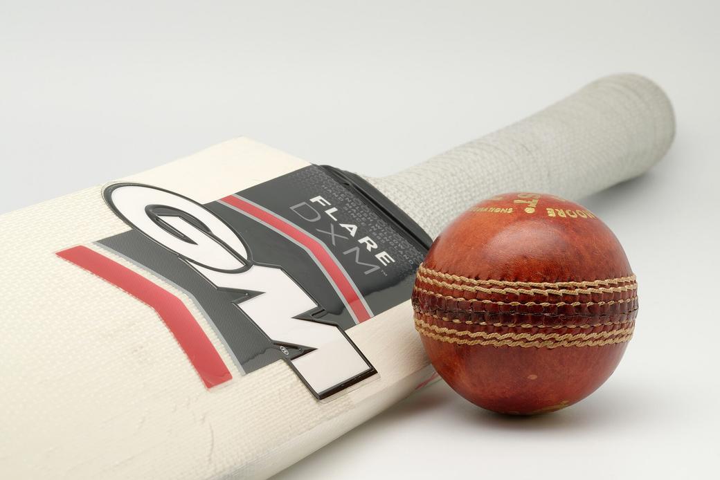A cream cricket bat with a dark Flare DXM label and a red leather cricket ball on a plain light background.
