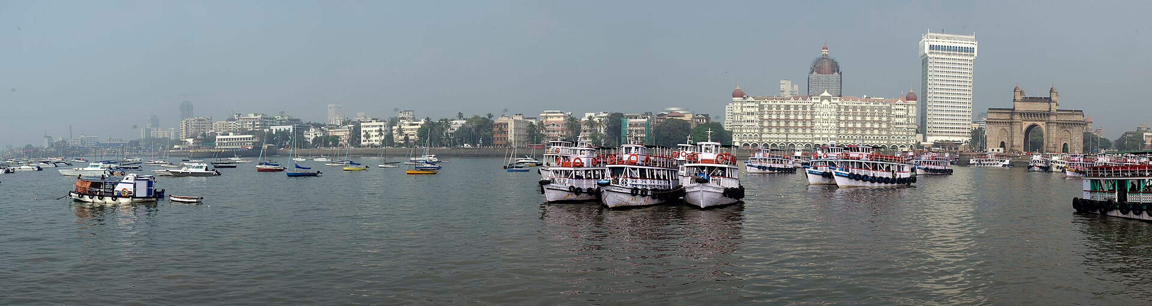Panoramic view of Mumbai's waterfront with ferry boats on hazy water, the Taj hotel and Gateway of India.